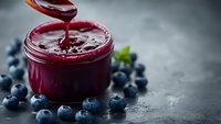 Homemade blueberry jam being poured from wooden spoon into glass jar with fresh berries scattered on dark stone surface for breakfast and cooking.