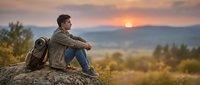 Male hiker with backpack sitting on rock at sunset, enjoying mountain landscape view during outdoor adventure and travel.
