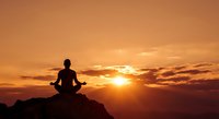 Person meditating in lotus position on mountain peak during golden sunset with dramatic orange sky and clouds for wellness and mindfulness concepts.