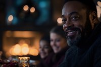 Smiling African American man with beard enjoying evening gathering, warm bokeh lights create cozy atmosphere as friends celebrate together at dinner party.