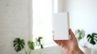 Person holding blank white box packaging in bright interior space with potted plants and natural window light for product display.