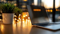 Laptop on wooden desk with decorative plant and warm fairy lights creating cozy evening atmosphere. Blurred city lights in background add ambiance to home office setup.