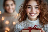 Young Caucasian woman with wavy brown hair and bright smile holding wrapped gift box with red ribbon, wearing cozy knit sweater against blurred holiday lights background.