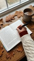 Person writing in open book at wooden desk with autumn leaves, warm beverage, and window view, capturing cozy fall season reading and journaling moments.