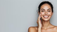 Young woman with glowing natural skin and bright white smile touching face gently against light gray background, showcasing skincare and dental aesthetics.