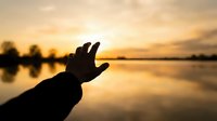 Silhouette of open hand reaching toward golden sunset over calm lake water with peaceful reflection and warm orange sky for meditation concepts.