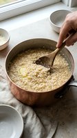 Cook stirring creamy risotto in copper pot with wooden spoon, preparing Italian rice dish in kitchen with natural light from window.