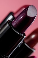 Dark burgundy lipstick with glitter sparkles on pink background, macro close-up shot showing luxurious texture and shimmer detail in cosmetic product.