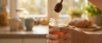 Honey dripping from wooden dipper into glass jar held by hand in warm kitchen setting with natural light and blurred background.