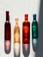 Colorful glass bottles in red, amber, orange, and teal arranged in row with dramatic sunlight creating geometric shadow patterns on white surface.