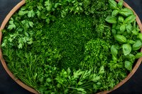 Fresh aromatic herbs and leafy greens arranged in wooden bowl on dark background, overhead view of parsley, dill, basil and other culinary herbs.