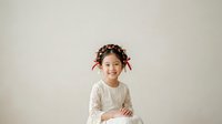 Young girl with braided hair and red ribbons wearing white lace long-sleeve shirt, smiling at camera against neutral background. Suitable for children's fashion, parenting, education, and lifestyle content.