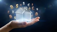 Hand holding transparent house model with floating currency symbols including dollar, euro, bitcoin, yen and pound representing real estate investment concept.