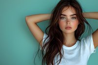 Young woman with blue eyes and long brown hair wearing white t-shirt poses against turquoise wall background, hands behind head in casual relaxed portrait.