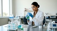 Scientist examining blue liquid sample in laboratory setting with test tubes and equipment on table.