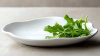 Fresh green arugula leaves served on modern white oval ceramic plate against neutral beige tablecloth background, minimalist food styling presentation.
