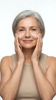 Mature Caucasian woman applying facial cream with gentle hands, showcasing anti-aging skincare routine and self-care for healthy glowing skin on white background.