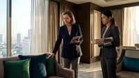 Two professional businesswomen in elegant dark suits discussing strategy in luxury office with city skyline view and modern interior design.