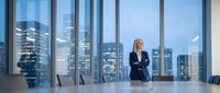 Confident blonde businesswoman standing in modern corporate boardroom with arms crossed overlooking illuminated city skyline through floor-to-ceiling windows at dusk.