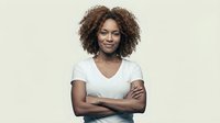 Young African American woman with curly hair smiling confidently with crossed arms wearing white v-neck shirt on neutral background for business portraits.
