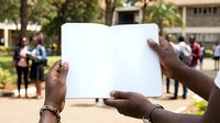 Person of African descent holding open blank white book or notebook with blurred community gathering in background, suitable for education, literacy, learning, and community development concepts.
