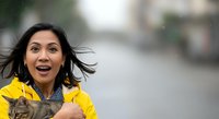 Young woman of South Asian descent wearing a bright yellow raincoat, holding a tabby cat while standing in rain on an urban street. Her expression shows joy and surprise as water droplets fall around her.
