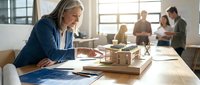 Mature female architect with gray hair reviewing building model and blueprints at wooden desk while diverse colleagues collaborate in background office space.