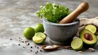 Fresh cilantro in stone mortar with wooden pestle, limes, avocado, salt and spices on rustic concrete surface for preparing guacamole or salsa.