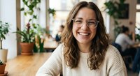 Young professional woman with glasses smiling warmly in modern office workspace with plants. Casual business attire for corporate lifestyle and workplace wellness.