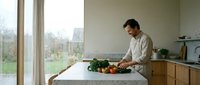 Young man preparing fresh vegetables on kitchen island in modern minimalist home with large windows overlooking garden during daytime cooking session.