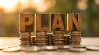 Wooden letter blocks spelling PLAN arranged on stacks of coins with warm sunlight background for financial planning and investment strategy concepts.