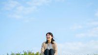 Young Asian woman wearing casual beige jacket and overalls standing outdoors against blue sky with clouds, looking upward with cheerful expression, suitable for lifestyle and fashion content.