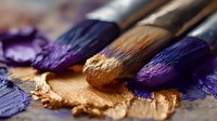 Professional artist paintbrushes with purple and gold paint on wooden palette, showcasing texture and color for fine art creation.