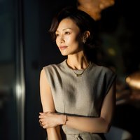 Professional Asian businesswoman in beige sleeveless top standing confidently with arms crossed in modern office environment with warm lighting.