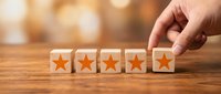 Hand placing wooden cube with orange star completing five-star rating row on wood table for customer feedback and quality assessment.
