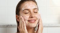 Young woman applying facial cleanser foam with hands in bathroom, enjoying skincare routine with eyes closed and peaceful expression for beauty wellness.