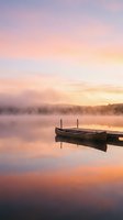 Moored wooden boat on calm misty lake at sunrise with soft pink and golden sky reflecting on still water, ideal for travel and nature backgrounds.