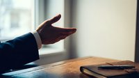 Businessman in dark suit gestures with open hand over wooden desk with notebook, representing communication, negotiation, or presentation in professional office setting.