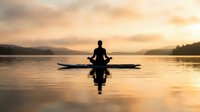 Silhouette of person meditating in lotus position on paddleboard at serene lake during golden hour sunset with mountain backdrop and calm water reflections.
