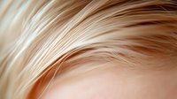 Soft focus macro shot of light blonde hair strands creating gentle waves and curves against blurred beige background, showing natural texture and shine.