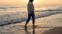 Woman walking barefoot on beach at sunset with golden light reflecting on wet sand and ocean waves creating peaceful coastal atmosphere.