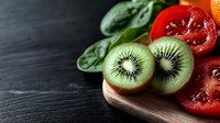 Fresh kiwi fruit halves with cucumber slices and red tomato pieces arranged on wooden cutting board against dark slate surface for healthy eating and food preparation.