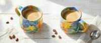 Two colorful ceramic mugs filled with creamy coffee on white wooden surface with scattered beans and spoon for beverage, morning routine, and café concepts.