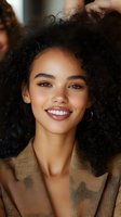 Young African American woman with curly hair smiling joyfully while touching her natural hairstyle, wearing beige blazer for beauty and lifestyle content.