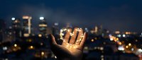 Young adult male hand holding glowing light bulb against blurred city skyline at night, representing innovation, ideas, and sustainable energy solutions for business and technology concepts.