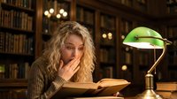 Young blonde woman reading book in vintage library with green banker lamp, studying late at night surrounded by wooden bookshelves and warm lighting atmosphere.