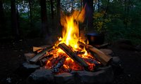Bright campfire with burning logs and flames in dark forest at dusk, stone fire pit with cooking pot, outdoor wilderness camping atmosphere.