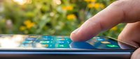 Finger touching smartphone screen with garden flowers blurred in background, representing mobile technology and outdoor connectivity.