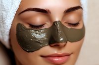 Young woman with clay face mask during spa treatment, wearing white towel on head, eyes closed with relaxed expression and natural makeup on lips.