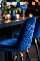 Luxurious navy blue velvet dining chair with quilted diamond pattern and gold legs against dark table setting with soft bokeh lights and floral decor.